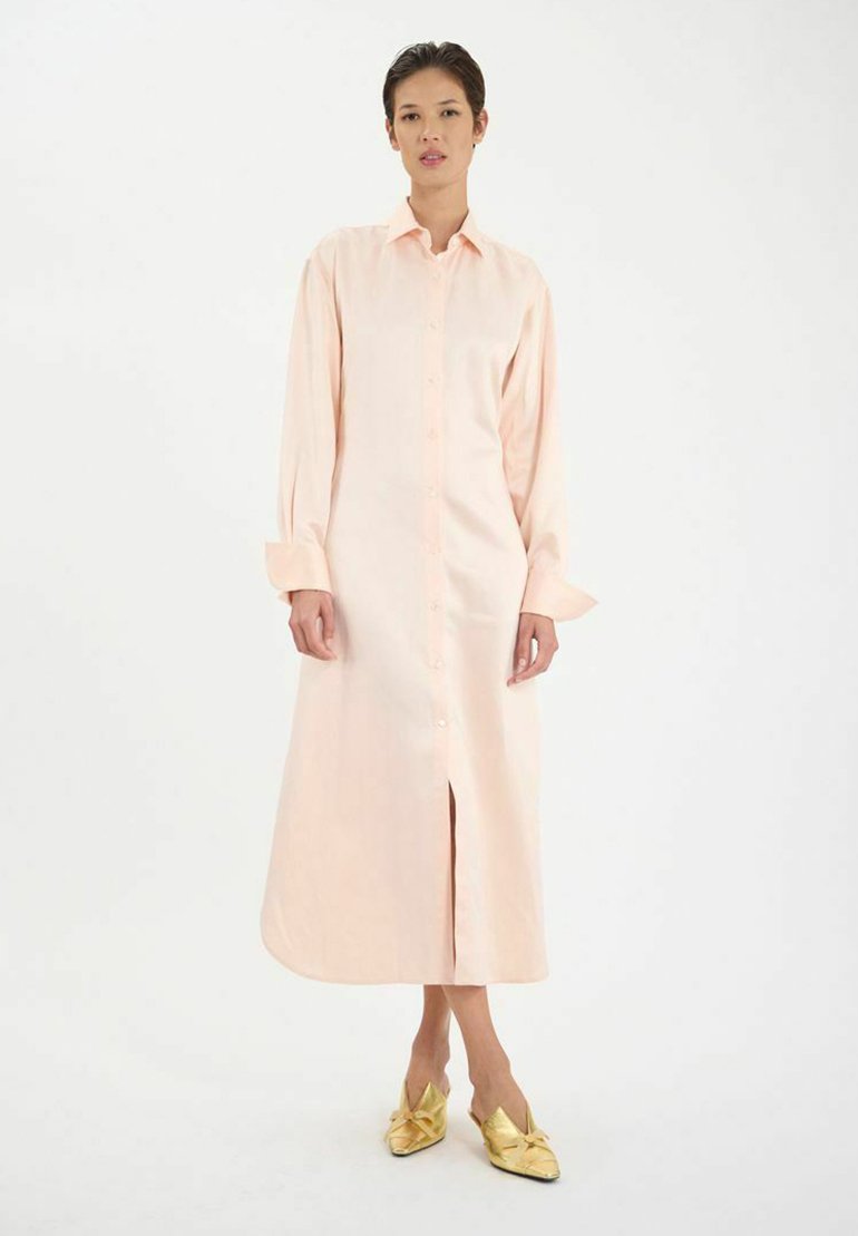 Robe boutonnière pêche clair avec des manches longues, présentant une fente avant et une coupe décontractée. Assortie à des chaussures à enfiler dorées et brillantes.