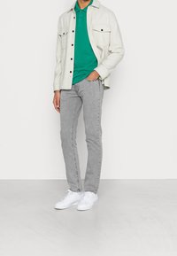 Jeans de mezclilla gris claro, camiseta polo verde y una camisa de botones beige con dos bolsillos frontales. Zapatillas blancas completan el conjunto.