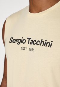 Ermeløs, lysegul bomullsskjorte med svart tekst som leser "Sergio Tacchini EST. 1966." Jevn tekstur, rund halslinning, ingen mønstre.