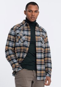 Geruit flanellen shirt met bruine, blauwe en gele patronen; heeft twee borstzakken en een gekraagde halslijn. Draagt over een donkere coltrui.