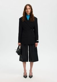 Γυναίκα με κυματιστά μαλλιά φορώντας μαύρο υπερμεγέθες σακάκι, μαύρο φαρδύ παντελόνι cropped, μπλε μπλούζα με φερμουάρ, κρατώντας μαύρη τσάντα χειρός και μαύρες μυτερές γόβες.