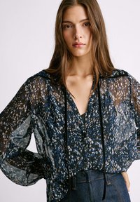 Blusa nera trasparente con stampa floreale blu e bianca, chiusura frontale a bottoni, maniche lunghe e dettagli con lacci neri.