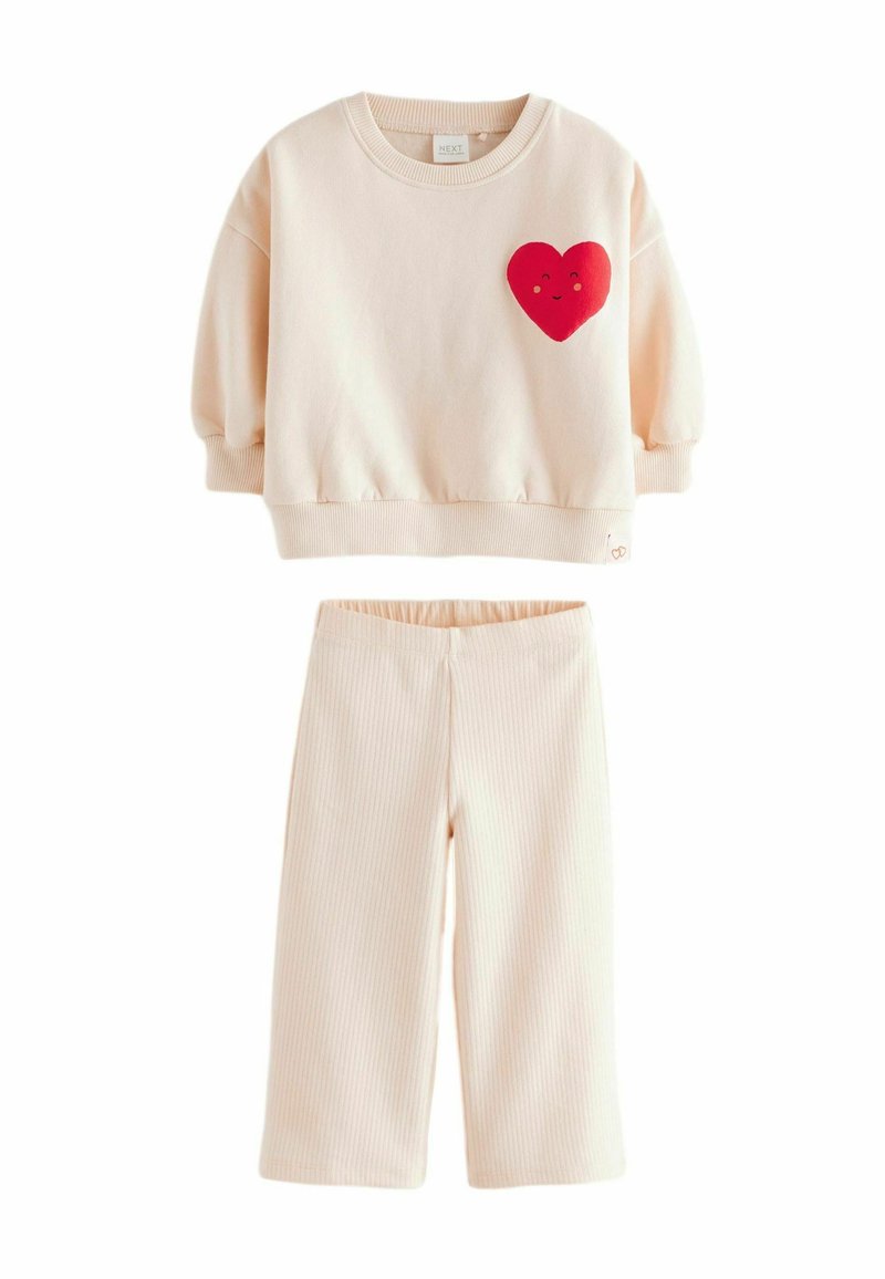 Sudadera corta beige con un parche de corazón rojo y una cara sonriente, combinada con pantalones beige de canalé. Tejido suave y corte relajado.