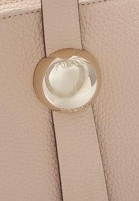 Svijetloružičasta kožna torba s glatkom teksturom. Ima okrugli zlatni detalj s ugraviranim natpisom "FURLA." Suptilni šavovi kao akcenti.