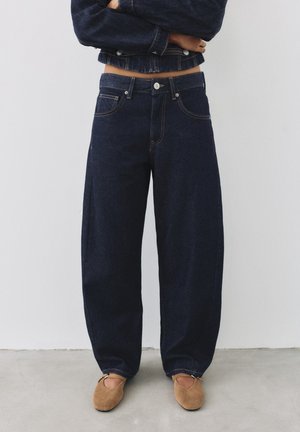 Personne debout, les bras croisés, portant un jean large foncé, une veste courte en denim et des chaussures plates marron sur un fond uni.