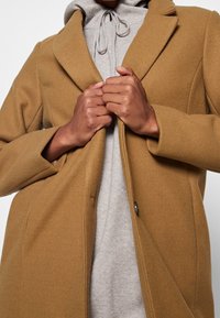 Brun kappa i ullblandning med skräddarsydd design, utrustad med en notchad krage och knappstängning, lager-på-lager över en ljusgrå huhoodie.