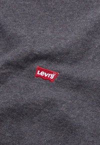 Tissu gris avec un logo Levi's rouge brodé en forme de badge emblématique, présentant une surface lisse et légèrement texturée.
