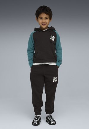 Ragazzo sorridente in piedi con le mani nelle tasche, indossando una felpa con cappuccio Puma nera e turchese, pantaloni della tuta abbinati, calzini bianchi e sneakers Puma nere.