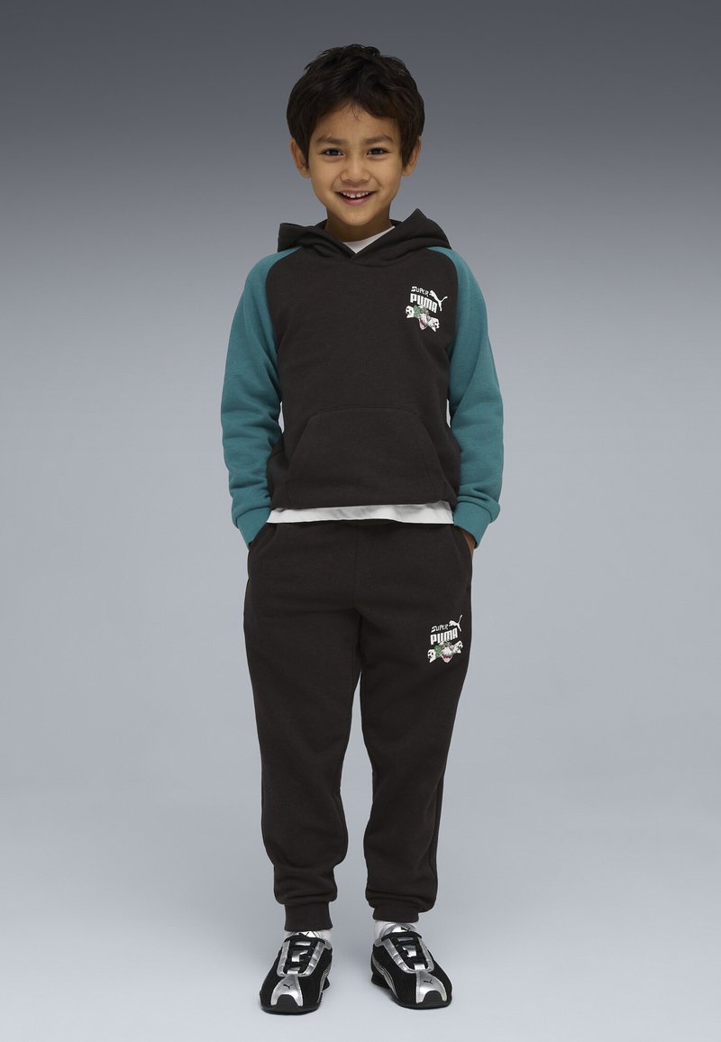 Glimlachende jongen die met de handen in zijn zakken staat, gekleed in een zwarte en turquoise Puma-hoodie, bijpassende joggingbroek, witte sokken en zwarte Puma-sneakers.