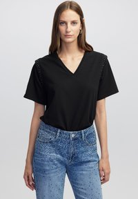 T-shirt nera a V con maniche corte e dettagli di borchie sulle spalle, abbinata a jeans blu decorati con strass.