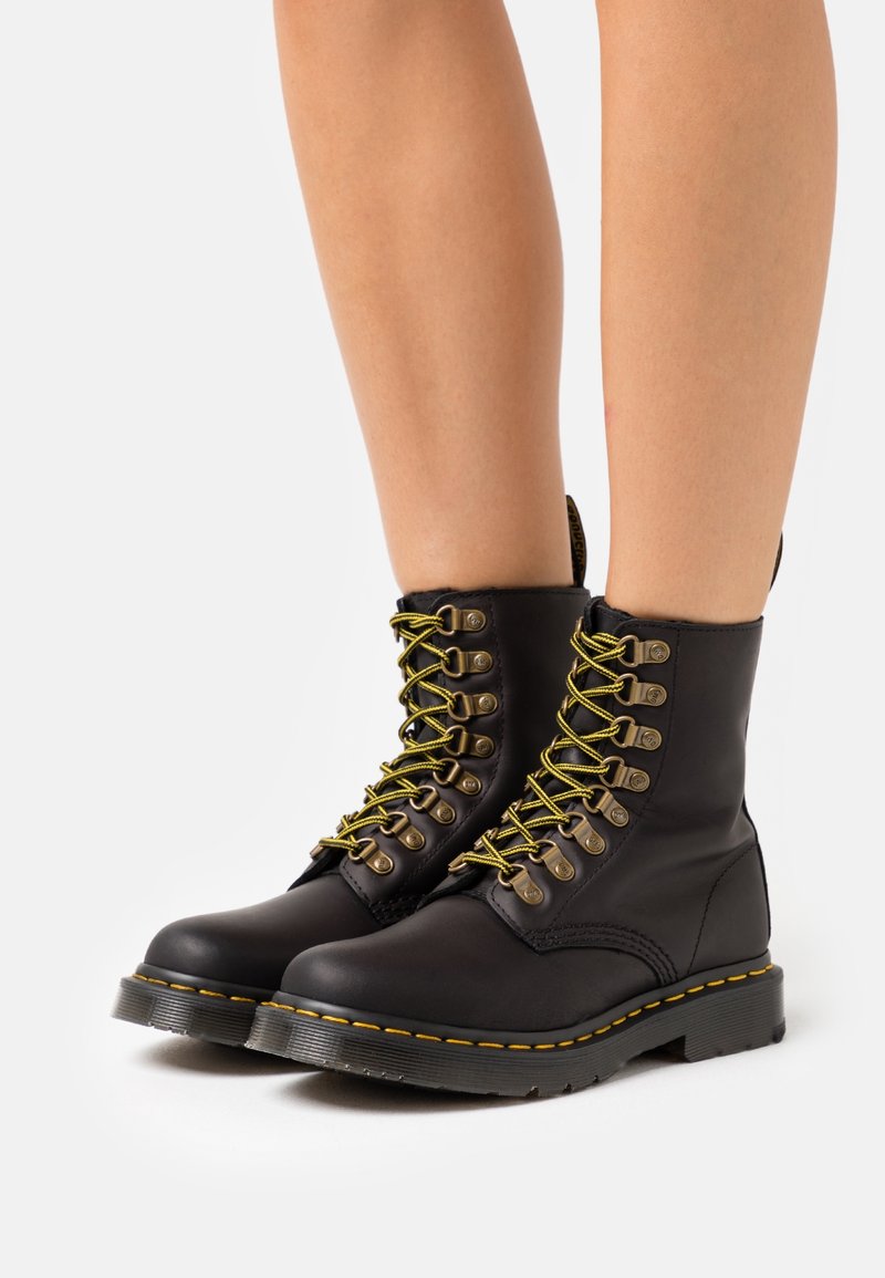 Botas de cuero negras hasta el tobillo con cordones amarillos y ojales dorados, que cuentan con una suela negra gruesa y costuras amarillas características.
