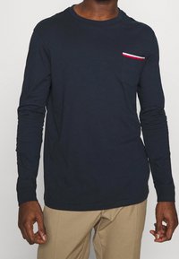 Mörkblå långärmad t-shirt i bomull med en bröstficka som har röd, vit och blå rand. Rundad hals med åtsittande ärmar.