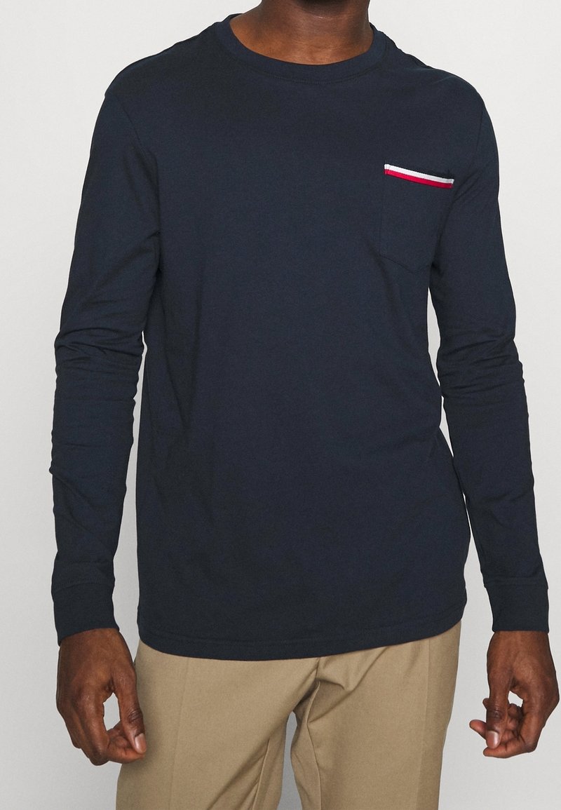 Mörkblå långärmad t-shirt i bomull med en bröstficka som har röd, vit och blå rand. Rundad hals med åtsittande ärmar.