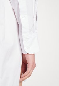 Chemise blanche à manches longues avec manchette à double bouton, tissu lisse et détail fendu sur le côté, avec une main naturelle posée sur l'ourlet.