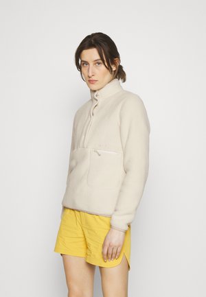 Beige fleece trøje med høj krave og halv lynlås, der har en frontlomme og struktureret stof. Kombineret med gule shorts.