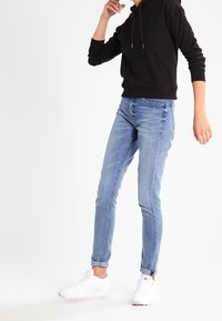 Jeans aderenti blu chiaro con vita media, abbinati a una felpa con cappuccio nera. Accentuati da scarpe sportive bianche. Texture liscia.