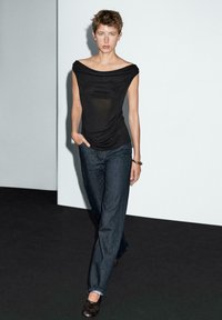 Svart draperad topp med vid halsringning, kombinerad med lösa mörkblå jeans. Modellen står mot en minimalistisk bakgrund.