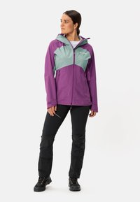Veste imperméable violette et vert clair avec une fermeture éclair, une capuche et deux poches, associée à un pantalon noir avec des panneaux texturés et des chaussures robustes.