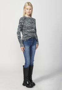 Pulover tricotat multicolor cu un model texturat, purtat cu blugi slim-fit albastri și cizme negre până la genunchi cu talpă groasă.