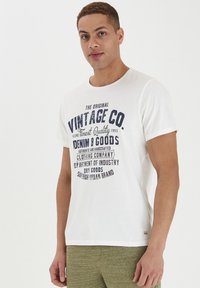 Bijela pamučna majica s plavim vintage grafičkim printom sprijeda. Okrugli izrez, kratke rukave, s malom oznakom branda na rubu.