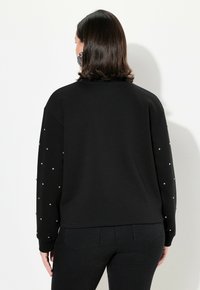 Felpa nera con maniche lunghe, caratterizzata da strass sparsi sulle maniche. Tessuto morbido e vestibilità rilassata. Indossata con jeans scuri.