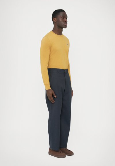 Camicia color senape a maniche lunghe con un piccolo logo, abbinata a pantaloni larghi blu scuro e sneakers marroni. Texture liscia su tutta la superficie.