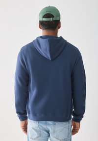 Marineblauwe trui met capuchon en een soepele textuur. Draagt men met lichtblauwe jeans; zichtbare achterkant bij de halslijn.