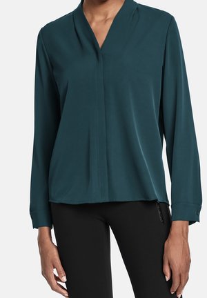 Blusa teal realizzata in tessuto liscio con scollo a V, maniche lunghe e un leggero orlo asimmetrico. Presenta un polsino con bottone e dettagli di cucitura minimi.