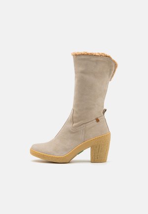 Botte mi-mollet en daim beige avec talon bloc en caoutchouc couleur cuir clair et doublure en mouton visible sur le bord supérieur.