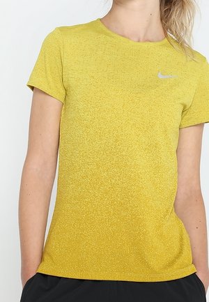 Kobieta ubrana w żółtą, krótkorękawową koszulkę sportową Nike z małym srebrnym logo Nike na piersi, ręce w czarnych kieszeniach spodni.