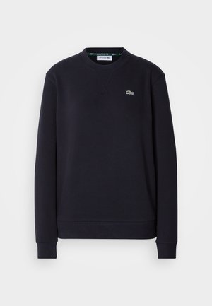 Enosivna črna dolgosleeve Lacoste majica s krožnim izrezom, z vezenim zelenim krokodilom na levi prsni strani, na beli podlagi.