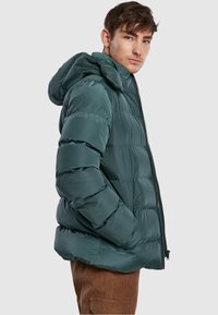 Tyrkysová péřová bunda se zipem a kapucí. Disponuje prošívaným designem, lesklou tkaninou a volným střihem se bočními kapsami.