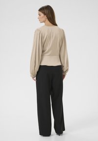 Blusa beige a maniche lunghe con maniche arricciate, texture liscia e vita, abbinata a pantaloni neri larghi. Design pulito e minimalista.