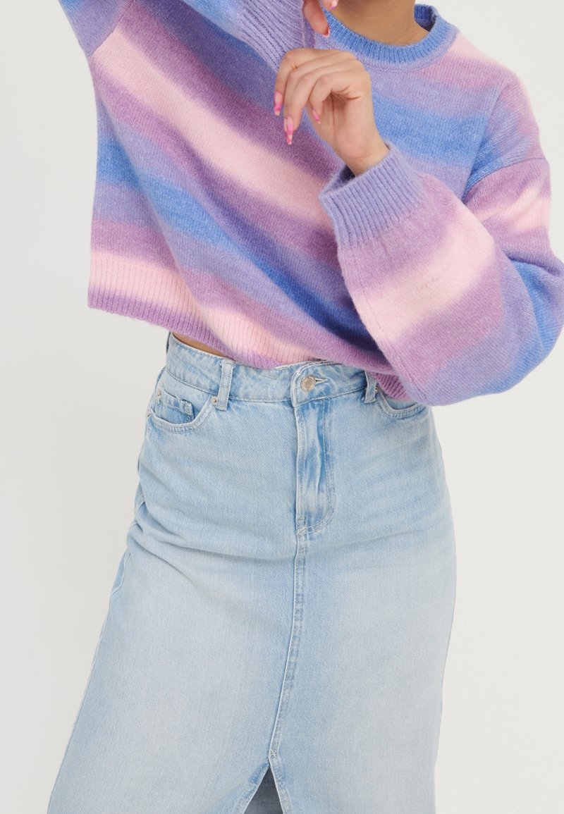 Personne portant un pull rayé pastel avec des nuances de violet, rose et bleu, associé à une jupe en jean bleu clair avec une fente à l'avant.
