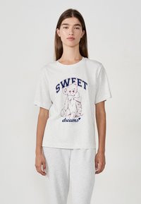 Jonge vrouw in een lichtgrijze T-shirt met een teddybeerafbeelding en de tekst "SWEET dreams", gecombineerd met bijpassende lichtgrijze broek.