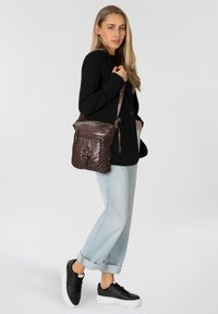 Borsa a tracolla in pelle marrone con texture intrecciata, dotata di chiusura con zip superiore e hardware decorativo. Abbinata a un abbigliamento casual.