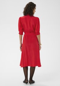 Robe midi rouge avec manches ballons, taille cintrée et jupe fluide. Fabriquée dans un tissu lisse, elle présente un léger éclat. Collants noirs et chaussures assorties.
