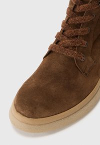 Brown suede ankle boot with a rounded toe, textured brown laces, and a light tan rubber sole. Features metal eyelets for laces.