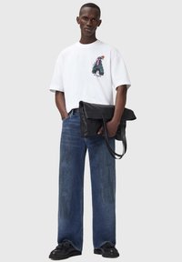 Hvid t-shirt med grafisk print på brystet, parret med løse blå denimjeans. Modellen holder en sort tekstureret taske med skulderremme.