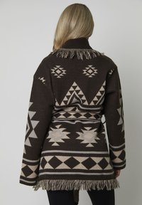 Een vrouw met blond haar staat met haar rug naar ons toe en draagt een donkerbruine gebreide cardigan met een patroon, afgewerkt met franjes en geometrische ontwerpen.