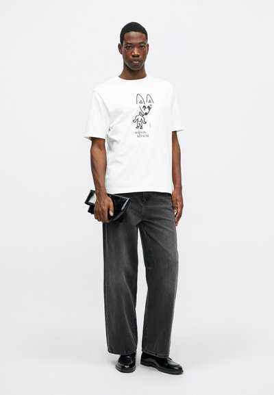 Mladý muž stojí v bílém tričku s potiskem, volných černých džínách, černých kožených botách a drží černou kabelku na jednobarevném pozadí.