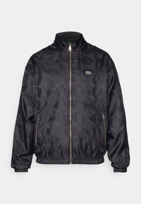 Čierna bomber bunda s geometrickým vzorom, zips na prednej strane, stojatý golier, elastické manžety a zelené logo Lacoste. Hladká, ľahká tkanina.