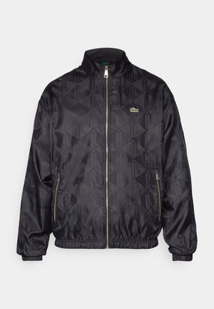 Černá bomber bunda s geometrickým vzorem, zipovým uzávěrem vpředu, stojatým límcem, elastickými manžetami a zeleným logem Lacoste. Hladká, lehká tkanina.