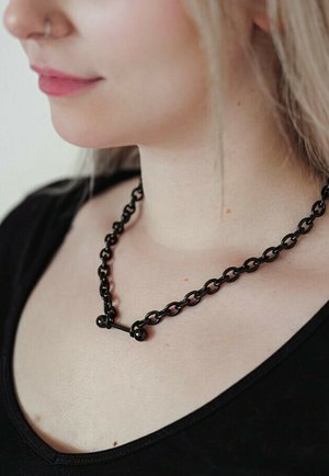 Frau trägt eine schwarze Halskette mit Gliedern und einem horizontalen Balkenanhänger, gekleidet in ein schwarzes V-Ausschnitt-Top, mit heller Haut und hellfarbenem Haar.