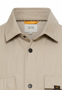 Chemise beige boutonnée avec col, deux poches poitrine, boutons noirs et une étiquette visible "camel active" à l'intérieur du col en taille large.