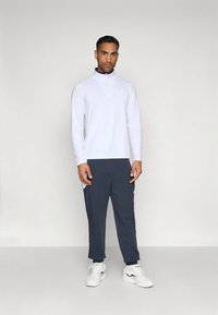 Muškarac stoji u bijelom dugom rukavu s patentnim zatvaračem, tamnosivim hlačama i bijelim sportskim cipelama, nasuprot običnoj svijetloj pozadini.