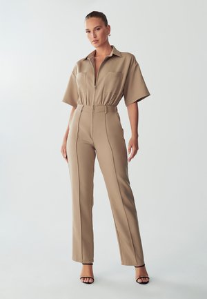 Tuta beige con maniche corte, zip frontale e vita elastica. Presenta due tasche sul petto e un design dritto e su misura.