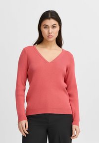 Koralinė V-formos megzta palaidinė su raukšlėta tekstūra ir ilgomis rankovėmis, derinama su juodais pantalonais. Medžiaga yra minkšta ir šiek tiek prigludusi.