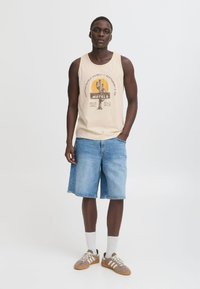 Top de tirantes de algodón beige con un estampado gráfico, pantalones cortos de mezclilla azul y zapatillas gris-marrón con rayas blancas y calcetines. Fondo liso.