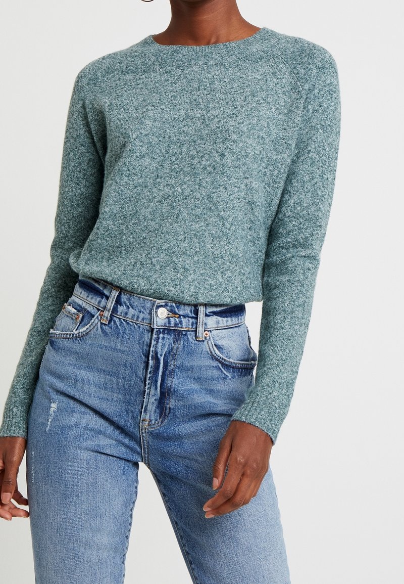 Femme portant un pull en maille à manches longues bleu sarcelle clair rentré dans un jean bleu délavé taille haute avec fermeture bouton et zip.